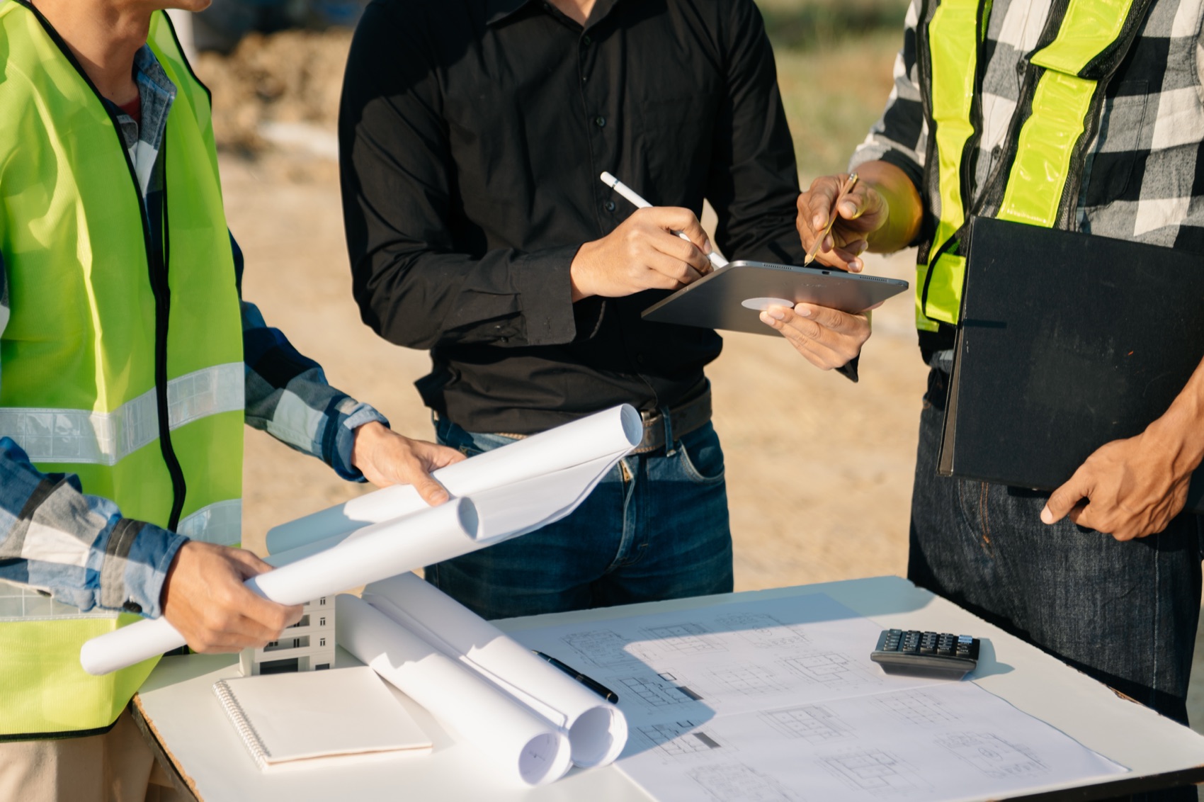 Three people around a site table with rolled drawings, a tablet and a floor plan.